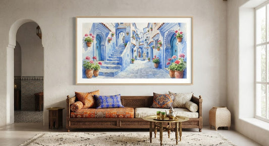 Grand tableau encadré représentant une aquarelle des ruelles bleues et fleuries de Chefchaouen, accroché au mur au-dessus d'un canapé en bois sculpté dans un salon marocain lumineux.