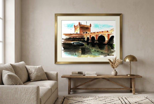 Tableau encadré représentant l'aquarelle La Sqala d'Essaouira, accroché au-dessus d'une console en bois dans un salon lumineux.